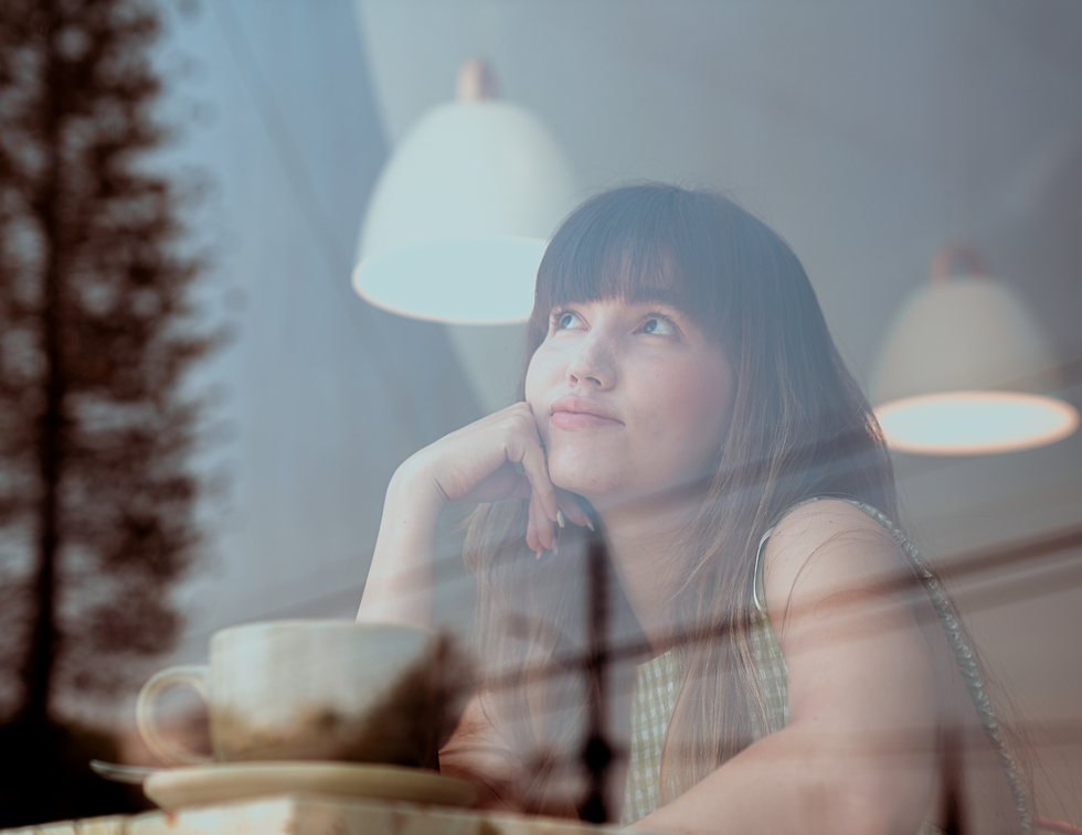 Woman in a cafe gazes thoughtfully out the window. Reflections of pendant lights and trees are visible. A cup sits in front of her.