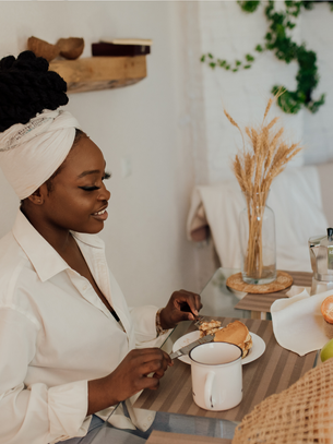 Woman with a headwrap enjoys breakfast at a table with fruit, a muffin, and a coffee pot. Cozy room with greenery and a vase of wheat.