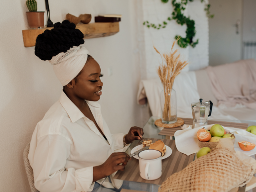 Woman with a headwrap enjoys breakfast at a table with fruit, a muffin, and a coffee pot. Cozy room with greenery and a vase of wheat.