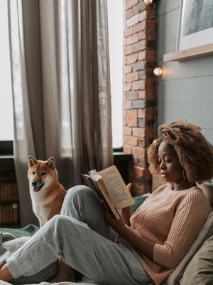 Woman reading a book on a cozy bed, beside a happy dog. Brick wall and curtains in the background. Warm, relaxing atmosphere.