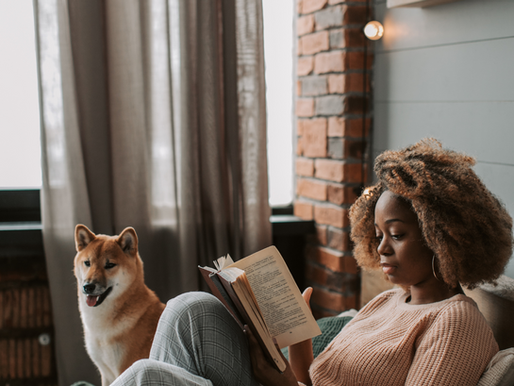 Woman reading a book on a cozy bed, beside a happy dog. Brick wall and curtains in the background. Warm, relaxing atmosphere.