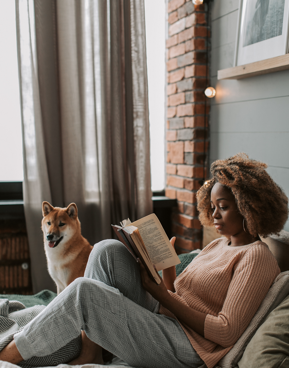 Woman reading a book on a cozy bed, beside a happy dog. Brick wall and curtains in the background. Warm, relaxing atmosphere.