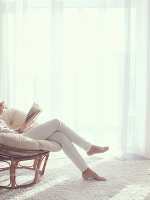 Woman in cozy chair reads book, sips coffee; soft natural light through sheer curtains. Calm, serene atmosphere in a bright room.