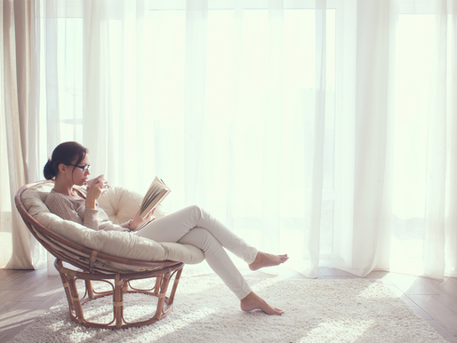 Woman in cozy chair reads book, sips coffee; soft natural light through sheer curtains. Calm, serene atmosphere in a bright room.