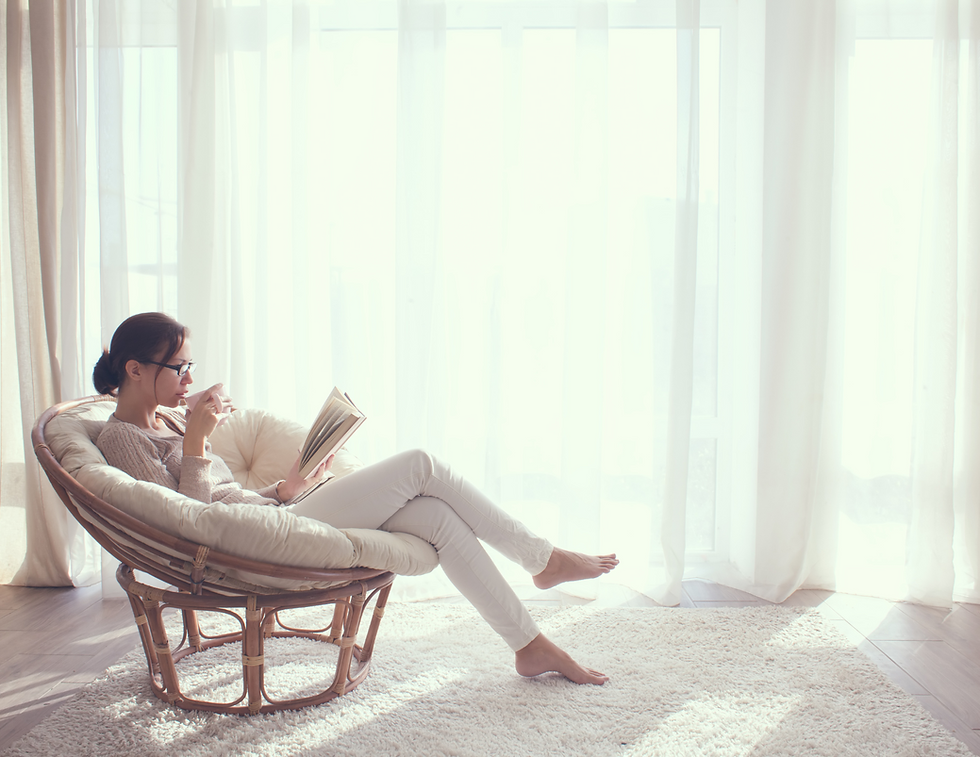 Woman in cozy chair reads book, sips coffee; soft natural light through sheer curtains. Calm, serene atmosphere in a bright room.