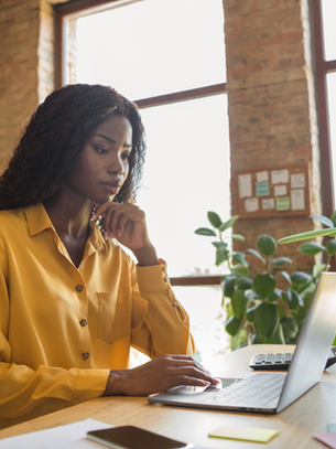 Woman in a yellow shirt works intently on a laptop at a desk with plants and stationery, in a sunlit office with brick walls and large windows.