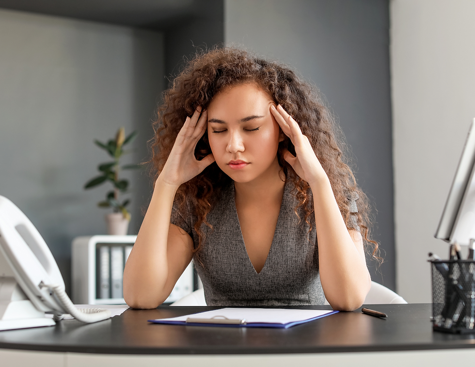 Woman in gray dress sitting at a desk, eyes closed, massaging temples. Office setting with a plant, papers, and phone. Appears stressed.