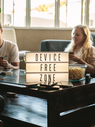 Four people play cards in a cozy living room. A sign reads "DEVICE FREE ZONE" on a table with popcorn. Relaxed, friendly mood.