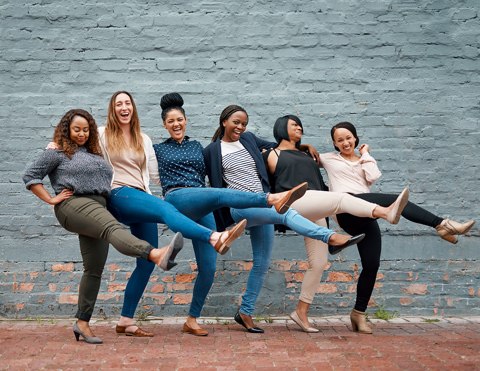 Six women smiling, kicking legs in unison, against a weathered blue brick wall. Casual attire, joyful mood, urban setting.