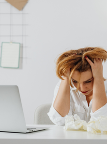 Alt text: Stressed woman sitting at desk with head in hands next to laptop representing mental fatigue vs burnout.