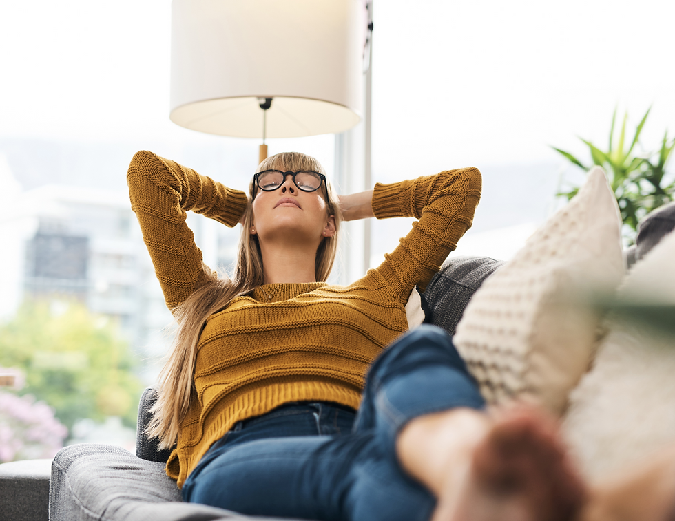 Woman in mustard sweater relaxes on sofa, eyes closed, hands behind head. Bright room with large window, plants, and cozy pillows.