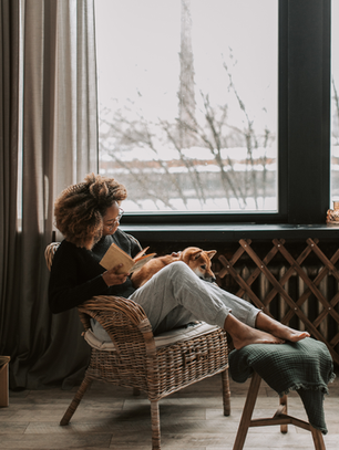 Black single mom practicing self-care guilt-free, relaxing in a cozy chair reading a book with her dog resting on her lap by a snowy window.