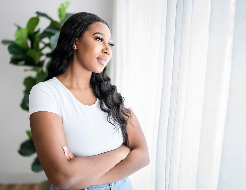 Woman in a white shirt stands with arms crossed by a bright window, gazing outside; green plant in the background.