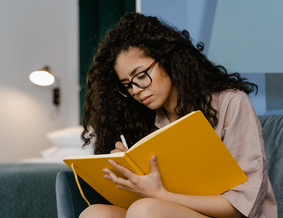 Woman with glasses writing in a yellow notebook, seated indoors in a cozy setting with a lamp in the background, looking focused.