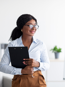 A confident woman standing in an office holding a clipboard, smiling with a calm and grounded presence, representing women embracing personal development and self-leadership.