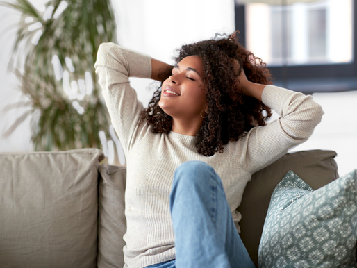 Woman relaxes on a grey couch, smiling with hands behind head. She's wearing a beige sweater and jeans. Green plant in the background.