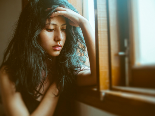 Woman with long hair leans on window, looking pensive. Sunlight filters through, creating a warm, contemplative mood. No visible text.