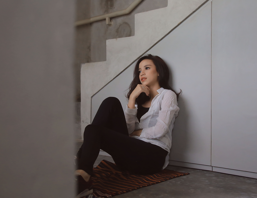 A woman in a white shirt sits pensively on the floor by a staircase, resting on a striped mat. The setting is minimalistic and calm.