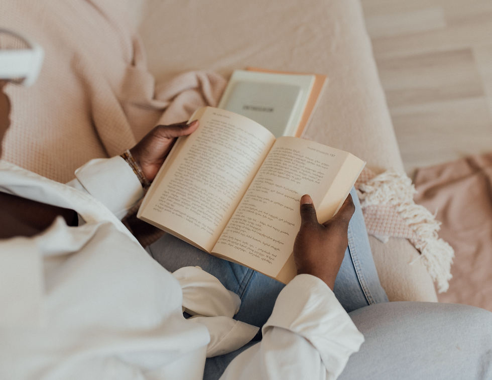Person in white shirt and jeans reading a book on a beige sofa. Another closed book nearby. Cozy, relaxed atmosphere with neutral tones.