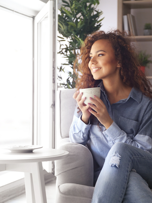 Woman sitting in a chair by a sunny window holding a coffee cup and smiling, representing a calm self-care routine for mental health