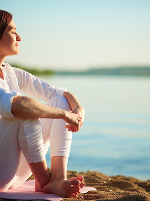 Woman in white sits on a beach, eyes closed, enjoying the sun. Calm water in the background, evoking a peaceful, serene mood.