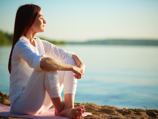 Woman in white sits on a beach, eyes closed, enjoying the sun. Calm water in the background, evoking a peaceful, serene mood.
