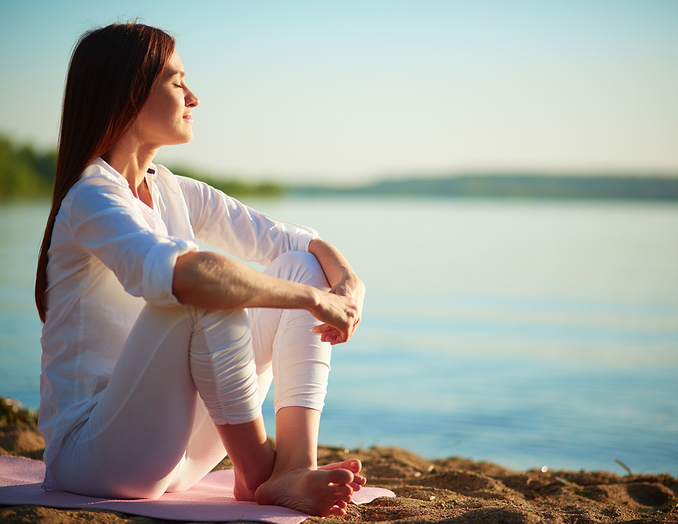 Woman in white sits on a beach, eyes closed, enjoying the sun. Calm water in the background, evoking a peaceful, serene mood.