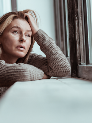 Woman in a gray sweater leans on a windowsill, gazing out with a pensive expression. Soft lighting, muted colors.