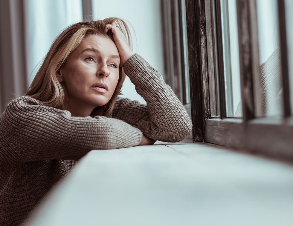 Woman in a gray sweater leans on a windowsill, gazing out with a pensive expression. Soft lighting, muted colors.