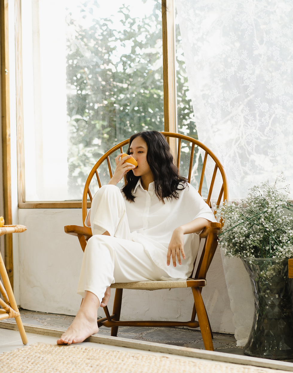 A woman in white clothes sits on a wooden chair, eating an orange. Soft light streams through large windows with lace curtains and greenery.