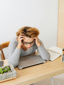 Overwhelmed woman experiencing warning signs of burnout in women, holding her head while sitting at a desk with a laptop