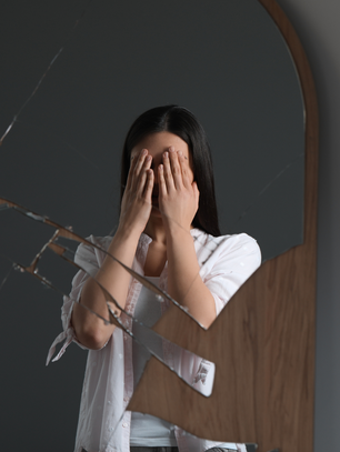 Woman covering her face in front of a broken mirror representing the signs of self-abandonment and emotional disconnection.