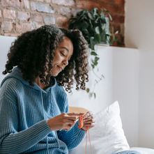 A woman sitting comfortably on a bed wearing a cozy blue sweater, smiling as she knits with orange yarn. The warm, relaxed scene reflects mindful self-care and calm creativity at home.