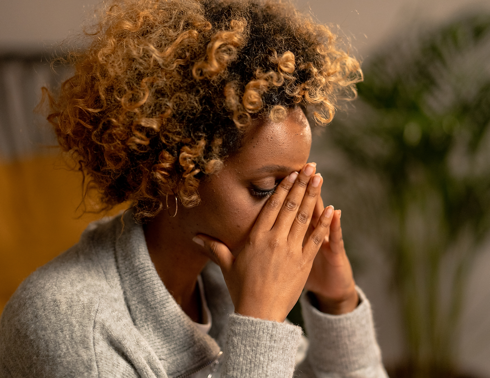 A person with curly hair sits with hands on face, appearing thoughtful. They're wearing a gray sweater in a soft-lit room with plants.