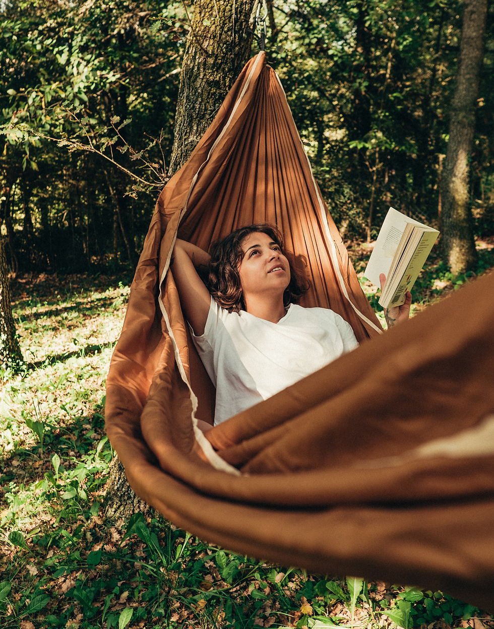 Person in a brown hammock, holding a book, looks content in a sunlit forest. Dappled light filters through leaves, creating a peaceful mood.