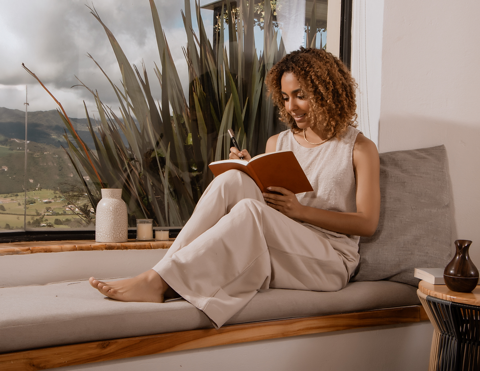 Woman with curly hair writing in a notebook, seated by a large window with a scenic view. She wears beige attire, exuding a calm mood.