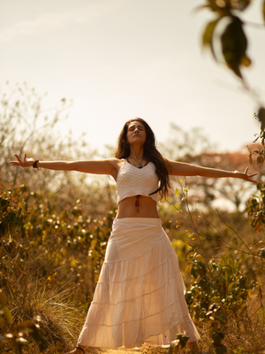 Woman in white top and skirt stands with arms outstretched in a sunny, leafy landscape. Serene expression, warm tones dominate.