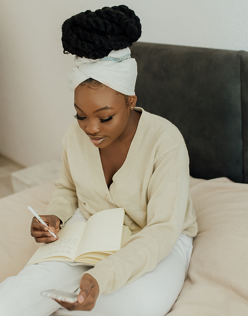 Young woman black woman wearing all white sitting on a bed with a pen and notebook in her hands.
