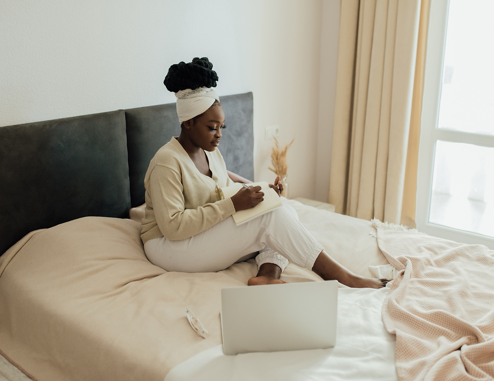 Woman sitting on a bed, writing in a notebook. She wears light clothing and a turban. A laptop and glasses lie nearby in a softly lit room.