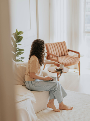 A woman with curly hair sitting barefoot on a couch with a laptop, practicing a 10-minute self-care ritual for overwhelmed women in a calm, bright living space.