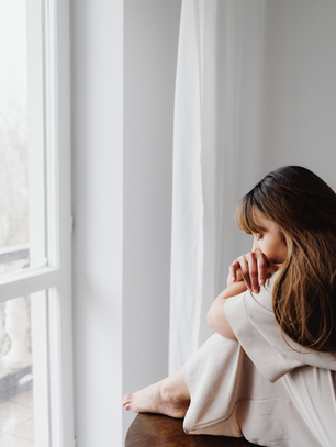Woman in a white dress sits pensively by a bright window, resting her head on her knees. The mood is contemplative and calm.