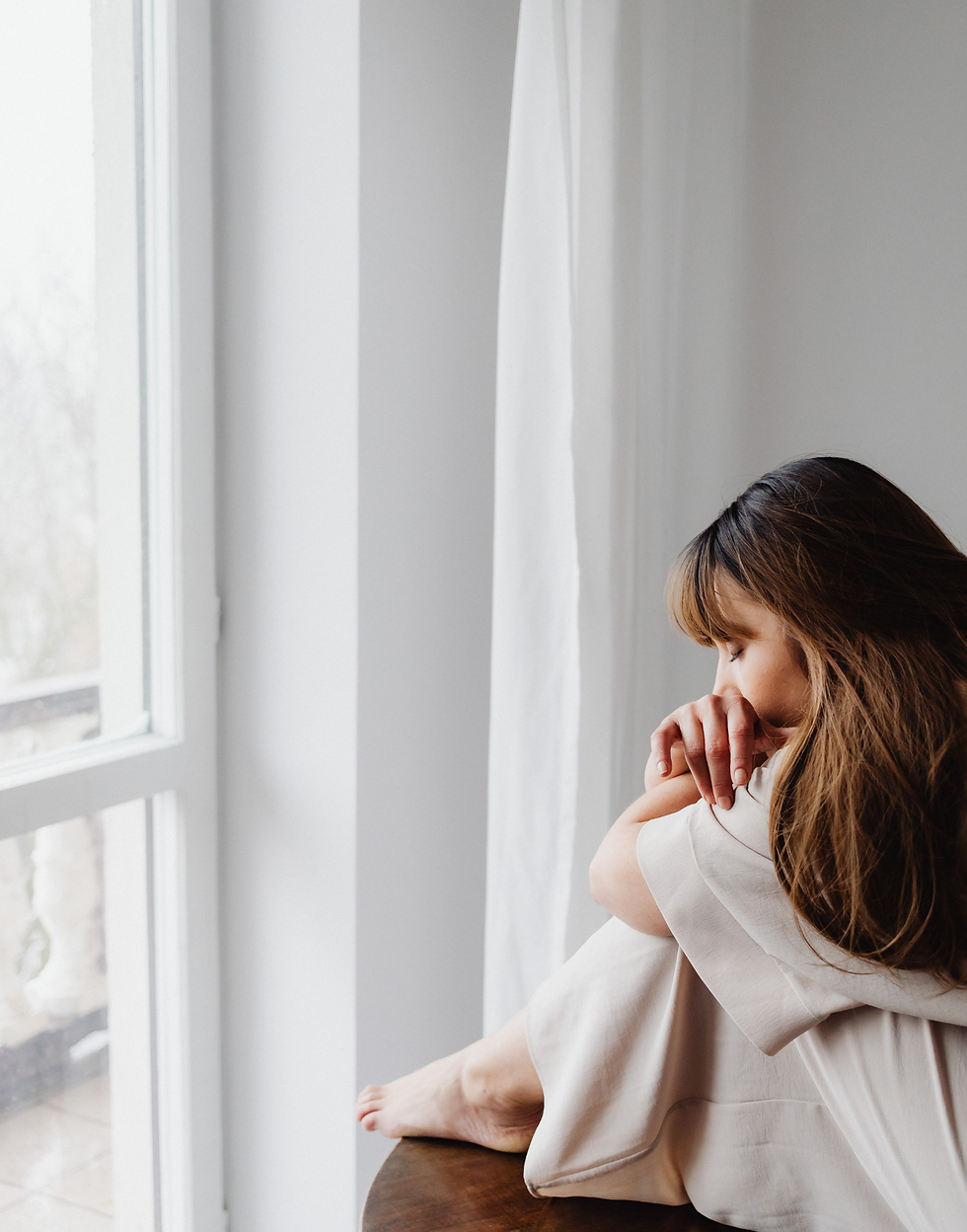 Woman in a white dress sits pensively by a bright window, resting her head on her knees. The mood is contemplative and calm.