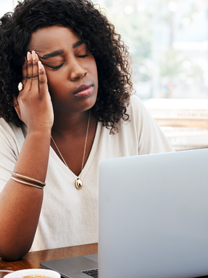 Woman with curly hair looks stressed, holding head, eyes closed, in front of laptop. Bright office with plants, books in background.