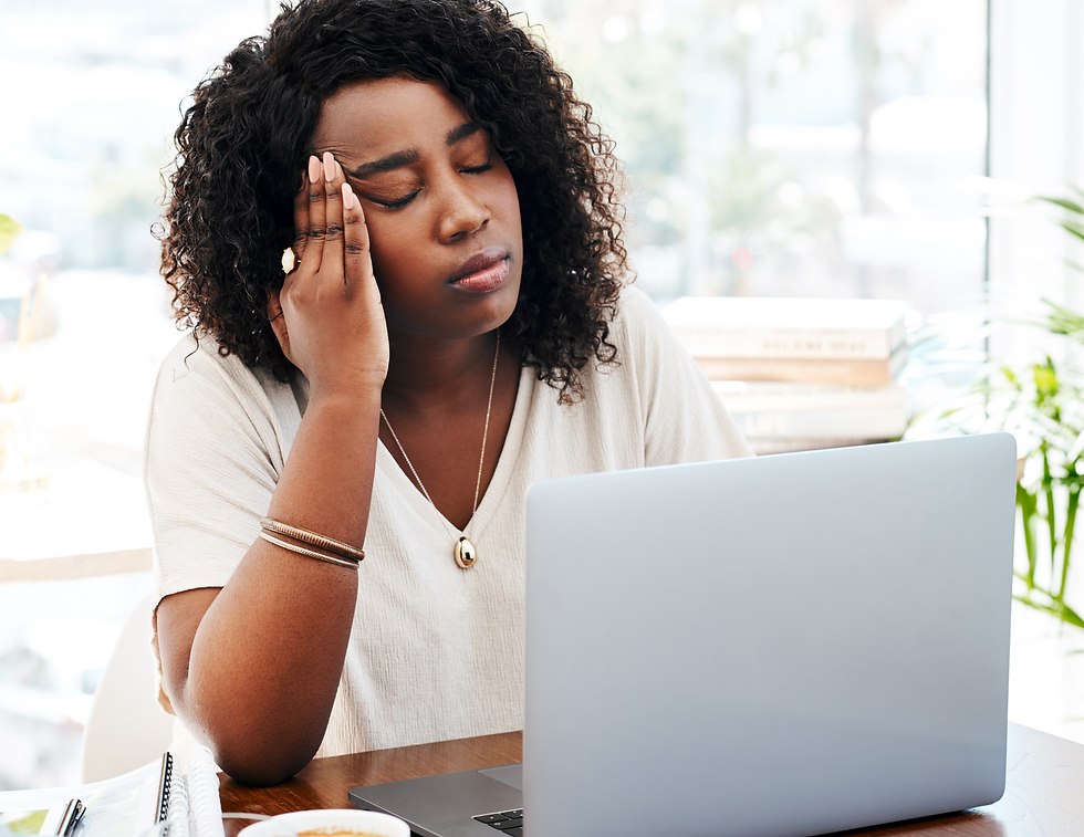 Woman with curly hair looks stressed, holding head, eyes closed, in front of laptop. Bright office with plants, books in background.