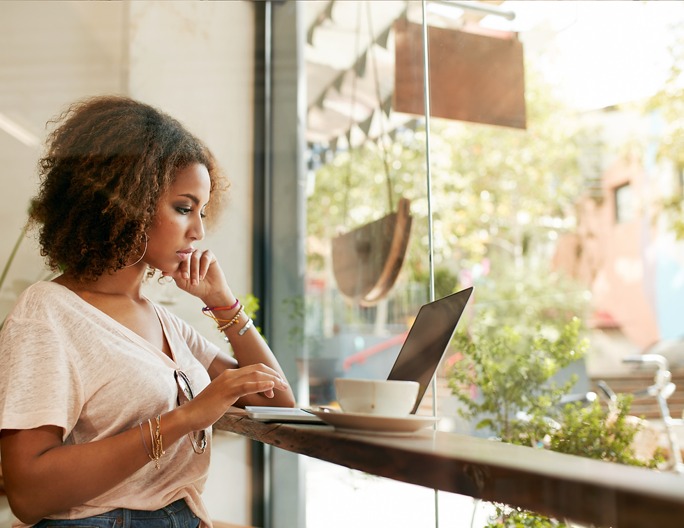 Woman in white shirt using laptop at a cafe window seat. Greenery outside, cup on table, focused expression. Bright, relaxed setting.