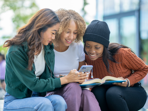 Three friends smiling and looking at a phone outdoors. One holds an open book. They're dressed in casual, colorful clothing. Bright, cheerful mood.