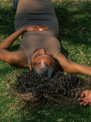 Woman in a brown dress lying on green grass, hands resting on her stomach. Sunlight highlights her face and hair. Calm, serene mood.