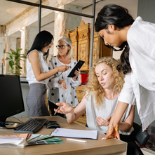 Four professional women collaborating in a modern office, reviewing documents at a desk and discussing work boundaries and communication during a team meeting.