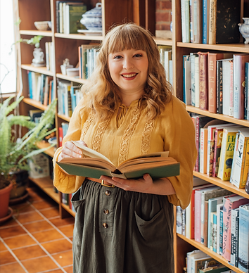 A photograph of Tess Carletta, a white, redheaded woman. She stands in a library holding an open book, dressed in a yellow blouse and green skirt. She's smiling.