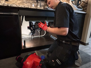 Person in black uniform fixes plumbing under a kitchen sink. Red toolbox nearby. Modern kitchen setting.